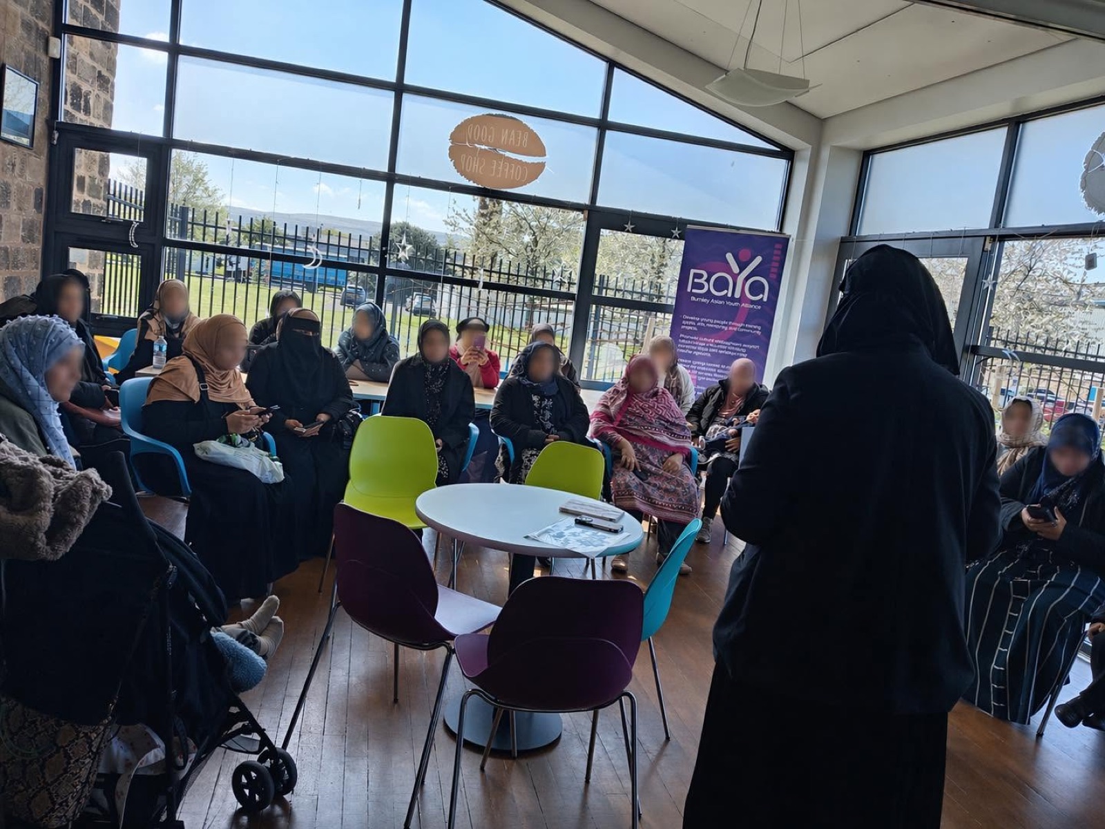 A speaker addressing a seated community group; participants’ faces are blurred in the supplied photograph.