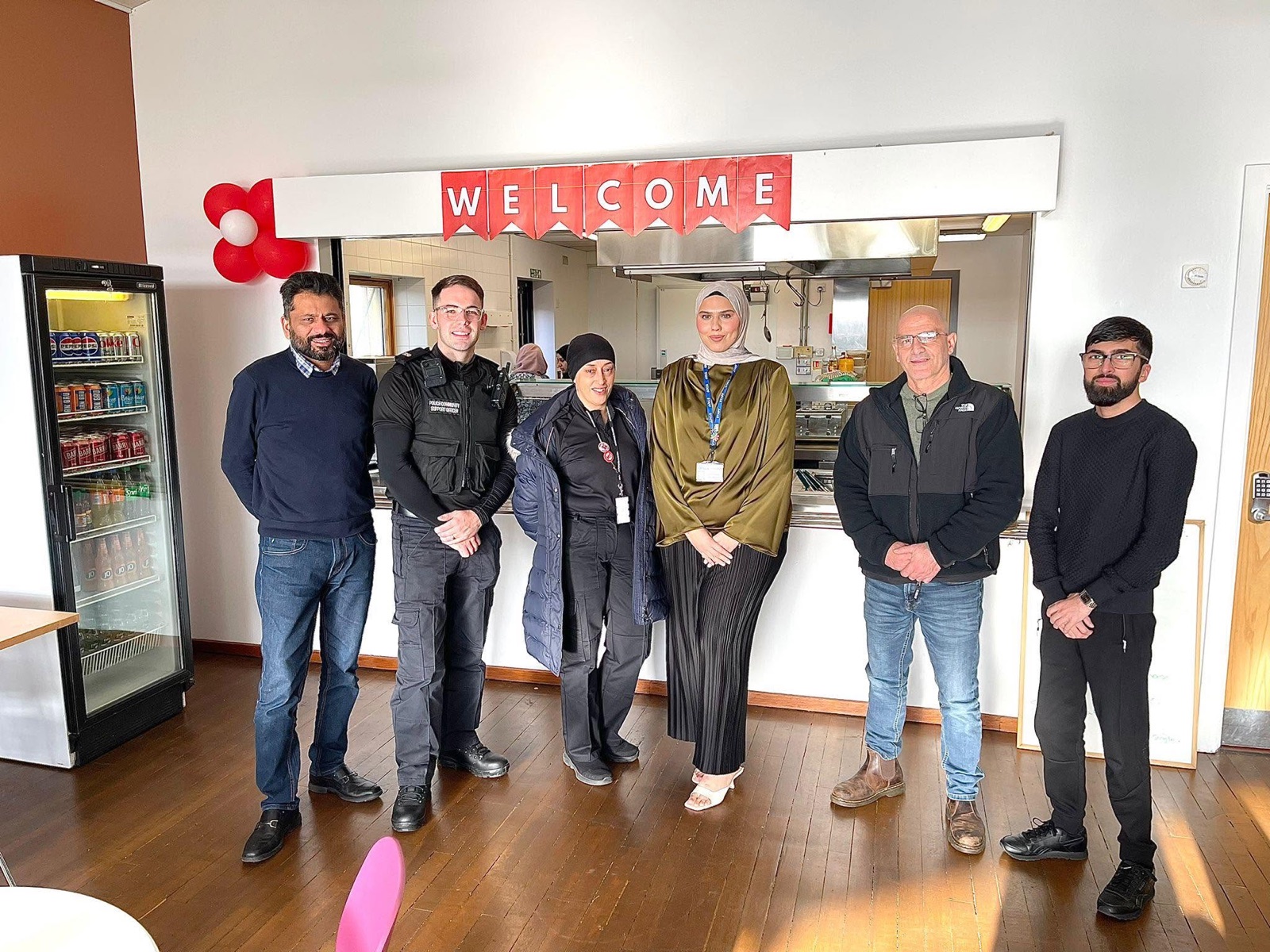 Six people standing together in a community cafe beneath a welcome banner.
