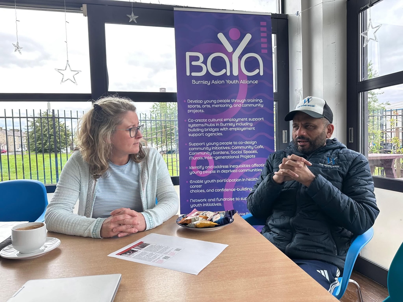 Two people talking at a table beside a Burnley Asian Youth Alliance banner.